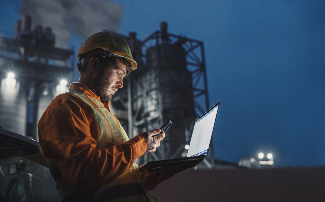 oil field worker on laptop and phone with refinery in background